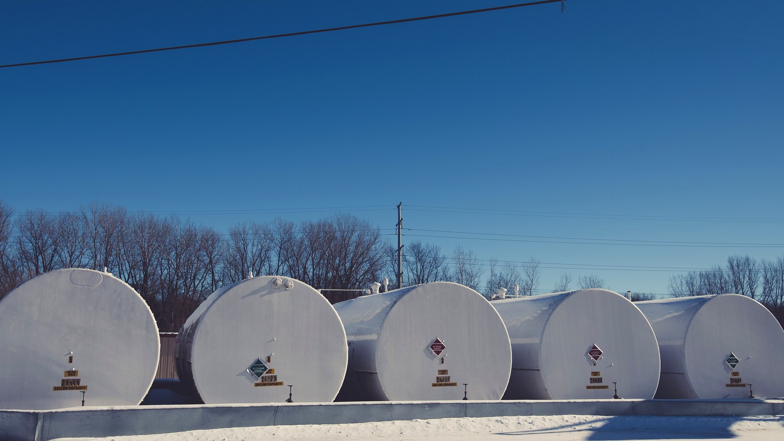Five white 20,000-gallon bulk fuel storage tanks in a row at a fuel distributor, with hazard placards reading #2 clear diesel and #2 dyed diesel, under a blue winter sky